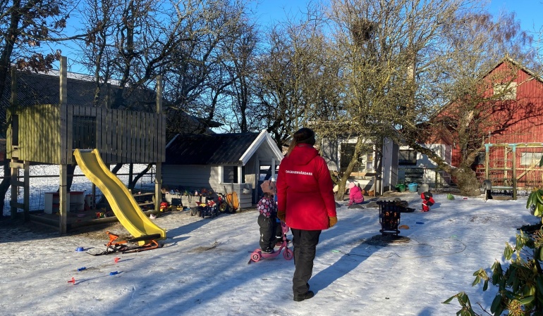 Dagbarnvårdare i röd jacka tittar på barn som leker utomhus.