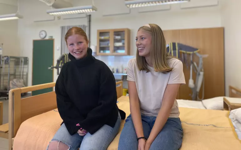 Enya och Ebba går vård och omsorgsprogrammet på gymnasiet, de sitter i en lektionssal på en sjukhussäng.