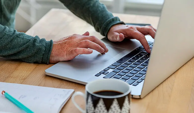 När bild på två händer som skriver på en silvrig laptop. I förgrunden syns en kaffe kopp och ett anteckningsblock.