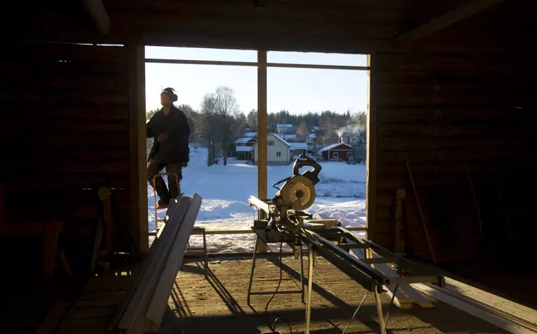 Bild tagen innifrån en lada, i öppningen står en såg, utanför ligger snö på marken och en man i arbetskläder och hörselkåpor går utanför