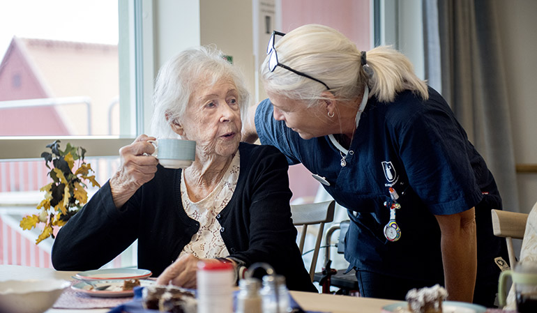 En äldre kvinna dricker kaffe medan hon pratar med en kvinnliga undersköterska.