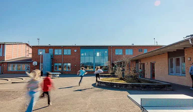 Åsa Gårds skolbyggnad. Byggnaden är i tegel med blåa spröjsade fönster. Fyra elever springer in mot byggnaden.