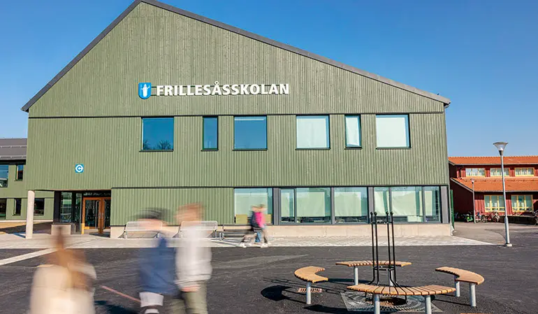 Frillesåsskolans gröna byggnad. Framför byggnaden står flera elever bredvid fyra bänkar.
