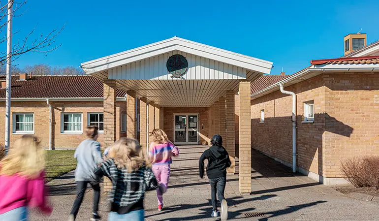 Fullriggaren Maleviks skolbyggnad. Byggnaden är i ljusbrunt tegel och flera elever syns springa mot entrén.