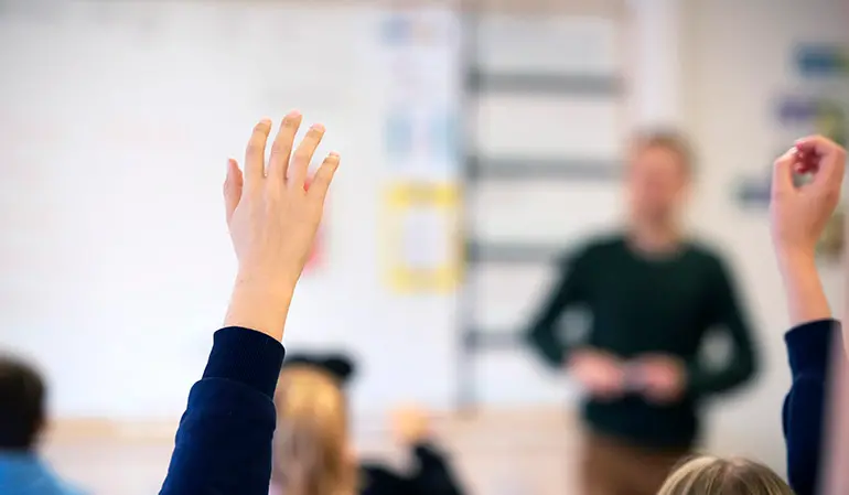 Elever som räcker upp händerna. I bakgrunden syn en lärare vid en whiteboardtavla.
