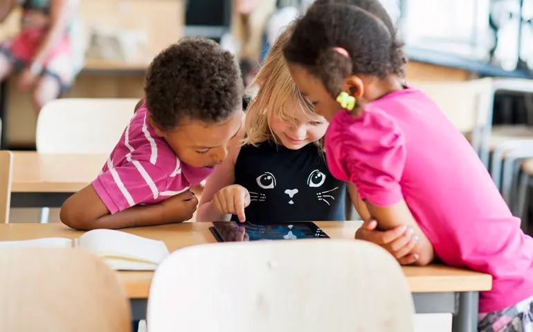 Tre barn tittar tillsammans på en läsplatta i ett klassrum.
