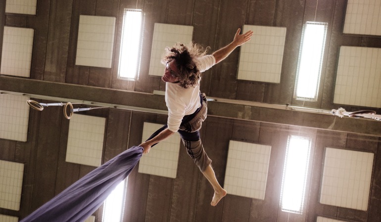 Man i vit skjorta och bruna byxor gör akrobatik i silkesband hängande från taket, bilden tagen underifrån 
