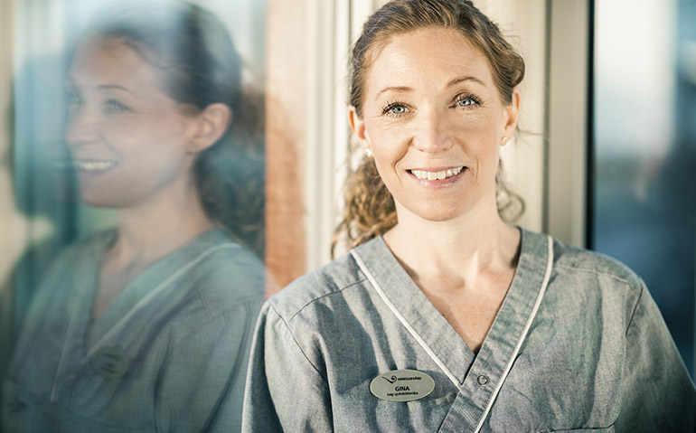 Porträttbild på en leende blond kvinna i undersköterskekläder och namnskylt med namnet Gina