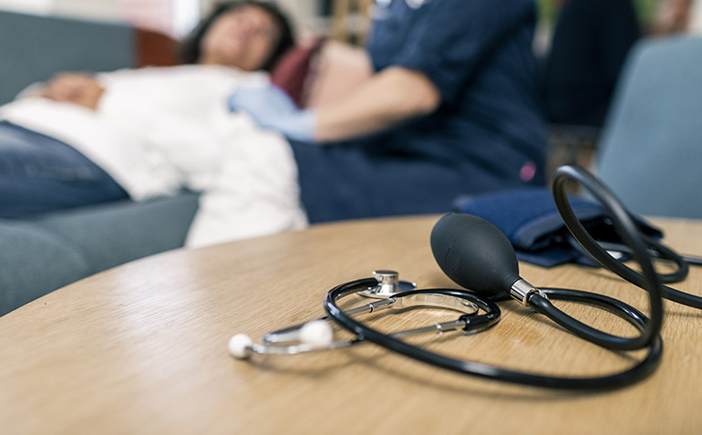 Stetoskop ligger på ett bord och i bakgrunden ligger en patient på en soffa.
