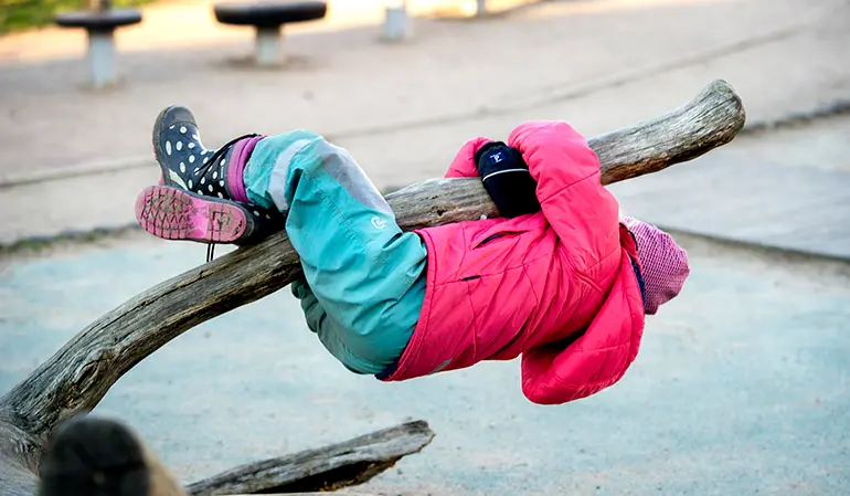 Barn i rosa jacka och turkosa byxor händer upp och ner från en låg trädgren.