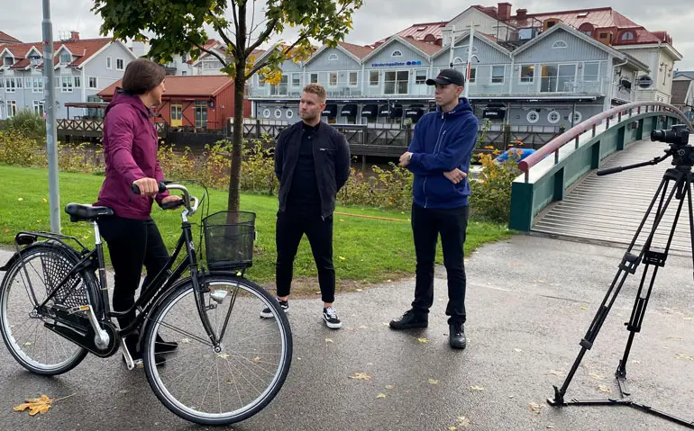 Elisabeth, Johan och Oscar står vid en bro. Elisabeth står med en cykel