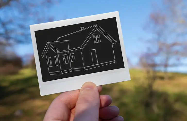 Närbild på en hand som håller upp en enkel ritning av ett hus, med blå himmel och en gräsmatta i bakgrunden 