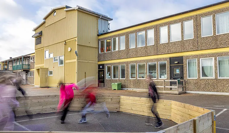 Gullregnsskolan bruna och gula byggnad. Framför byggnaden leker flera elever i en rink.