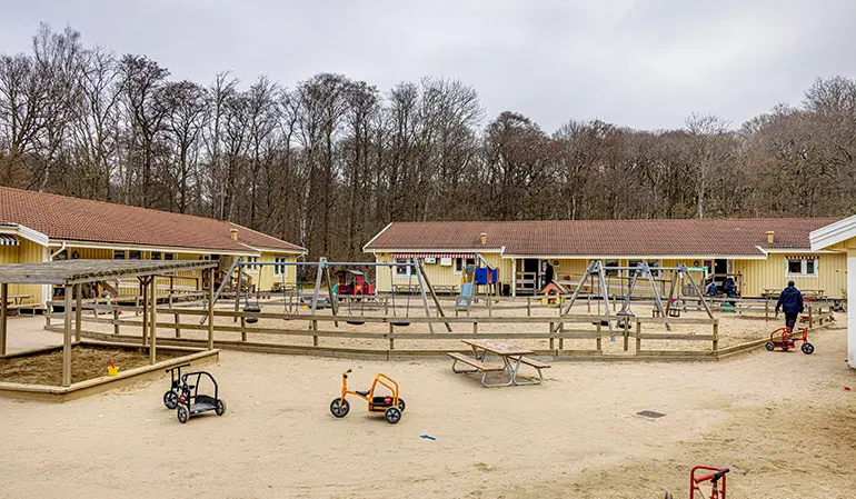 Förskolans två gula byggnader och gård med sandlåda och gungor.